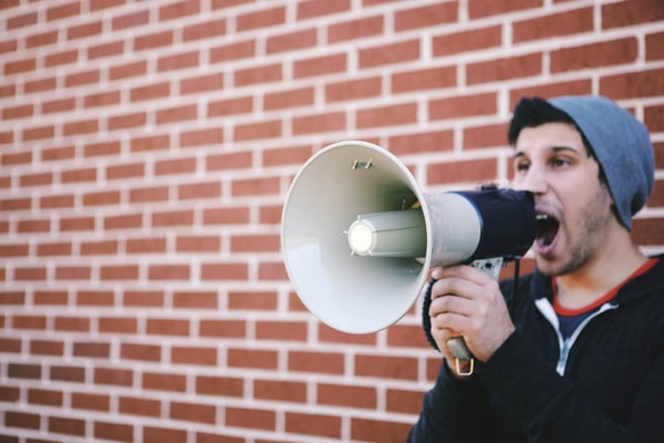 Young man in a beanie shouting into a megaphone in front of a brick wall