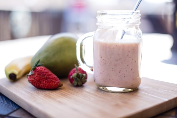 Pink fruit smoothie in a mason jar mug next to strawberries, a mango and a banana on a cutting board