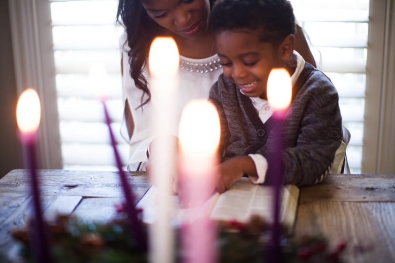 Smiling woman and young boy reading a book together at a wooden table behind lit Advent candles