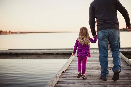 Man holding a young girl's hand as they walk along a wooden dock beside a lake