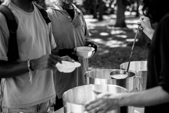 Person ladling soup from large pots into bowls held by people waiting in line outdoors, in black and white