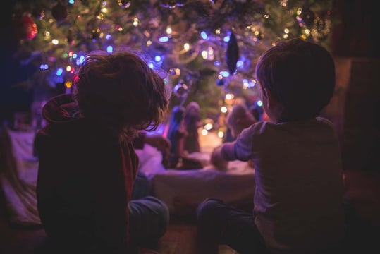Two children sitting by a lit Christmas tree, looking at a nativity scene