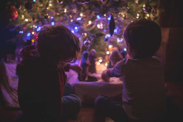 Two children sitting by a lit Christmas tree, looking at a nativity scene