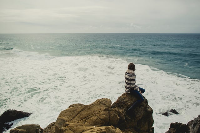 A woman sitting on the edge of a boulder overlooking the ocean.