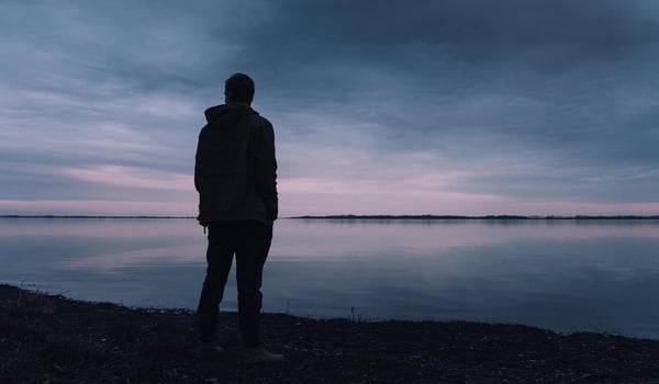 Silhouette of a man standing alone on a shoreline at twilight, looking out over calm water