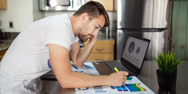 Man leaning over a kitchen counter with a hand to his head, highlighting printed charts beside a laptop
