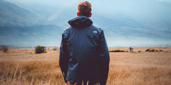 Man in a rain jacket, seen from behind, looking across a golden field toward misty mountains