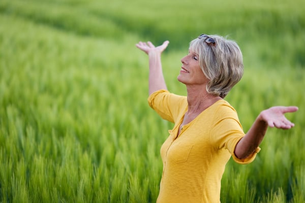Older woman with eyes closed and arms outstretched smiles in a green wheat field