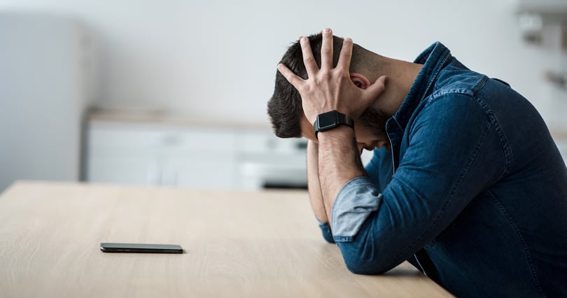 Man at a table holds his head in his hands beside his smartphone