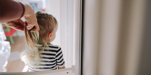 An adult's hands braid the long blond hair of a young girl in a striped shirt, seen from behind