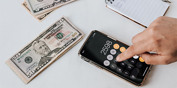 Hand using a phone calculator beside stacks of $50 bills and a spiral notepad