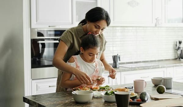 Woman standing behind a young girl, helping her prepare food at a kitchen counter