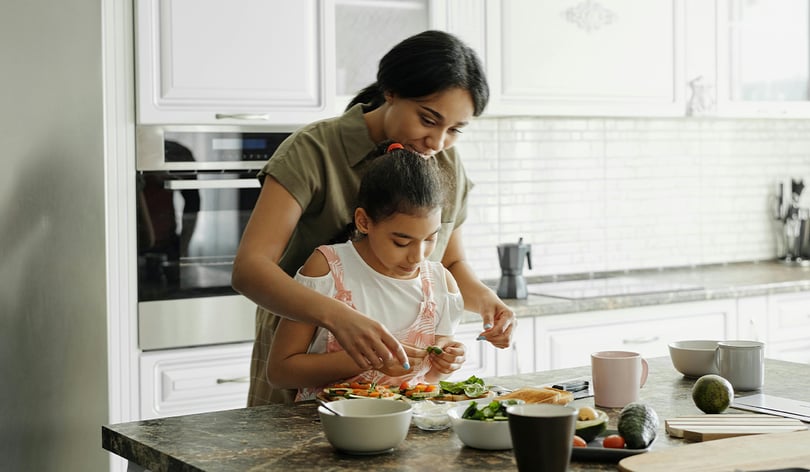 Woman standing behind a young girl, helping her prepare food at a kitchen counter