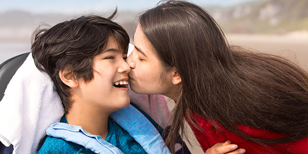 Young woman kissing the cheek of a laughing boy seated in a wheelchair at the beach