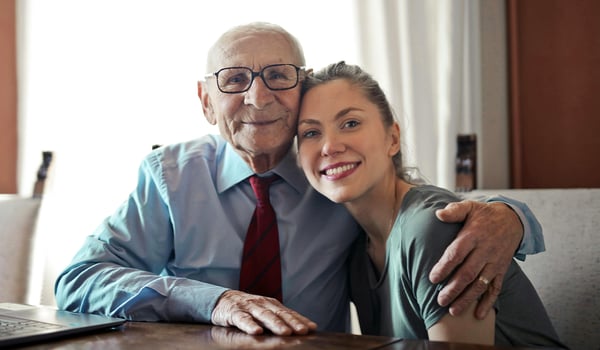 Older man in glasses and a tie smiling with his arm around a young woman leaning against him