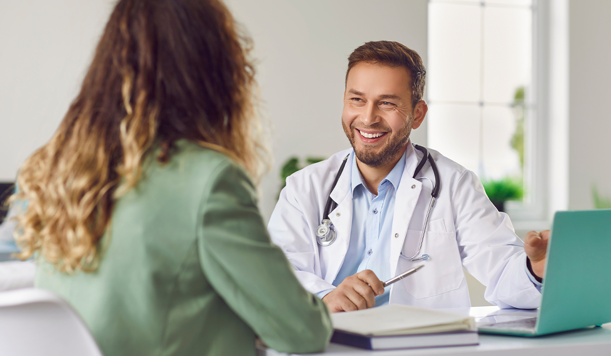 Medical consultation showing a healthcare professional at a desk with a laptop speaking with a seated patient.