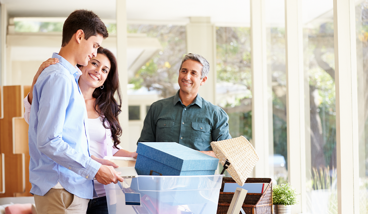 Teen boy packs personal items into storage bins and boxes indoors while preparing to leave for college while his parents look at him and smile.
