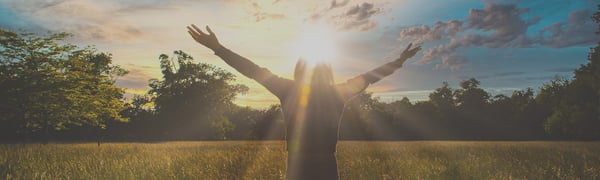 Person standing in a grassy field with arms raised wide toward the sun