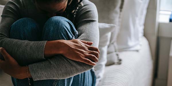 Person in a gray sweater and jeans sits curled up hugging their knees, face hidden, near a bright window