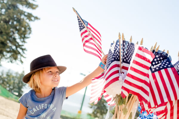Smiling child in a brown hat reaching toward a bundle of small American flags on a sunny day