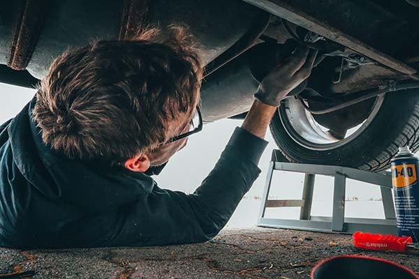 Man in glasses lying under a car on a jack stand, working on the wheel with a gloved hand
