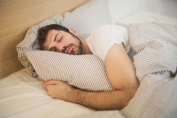 Bearded man in a white T-shirt sleeps soundly, hugging a striped pillow under a matching duvet