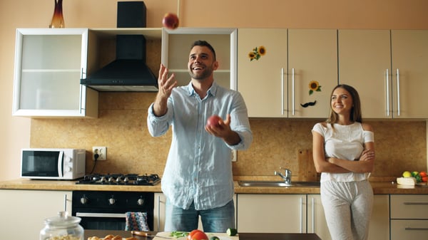 Man juggles apples in a bright kitchen as a smiling woman watches with her arms crossed