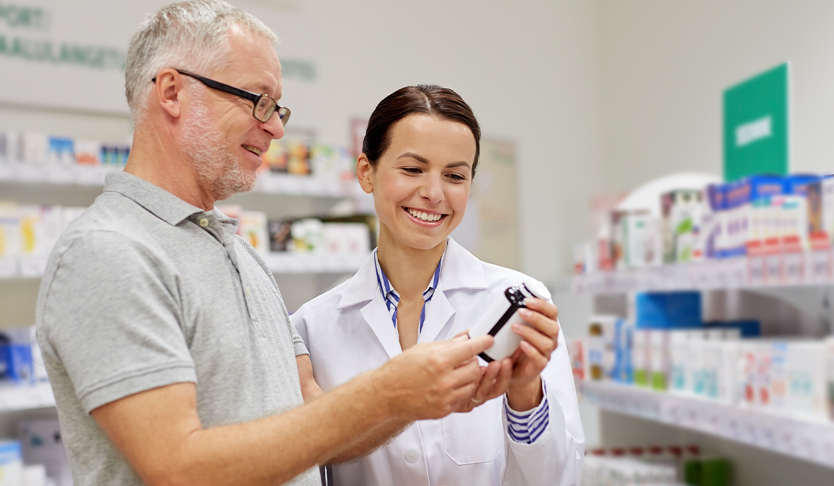 Two people stand in a pharmacy aisle looking at the label on a small bottle, with product shelves behind them.