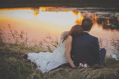 Bride and groom sitting close together on a grassy lakeshore at sunset