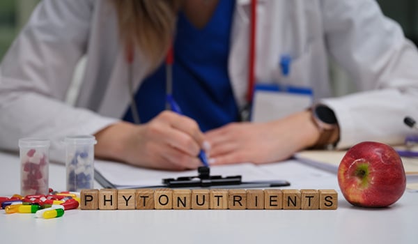 Wooden letter blocks spelling “phytonutrients” beside an apple and capsules as a doctor takes notes