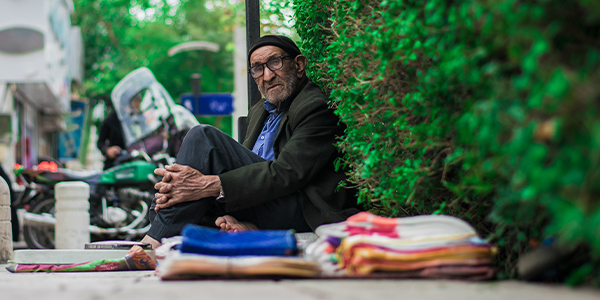 Older man in a cap and glasses sitting on a sidewalk against a hedge beside stacks of folded cloth