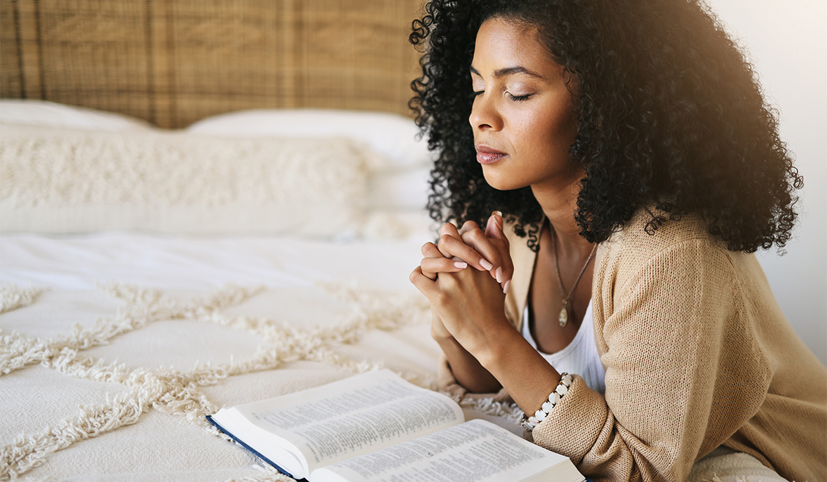 Person kneels beside a bed with clasped hands and an open book resting on the bedding in a quiet room.
