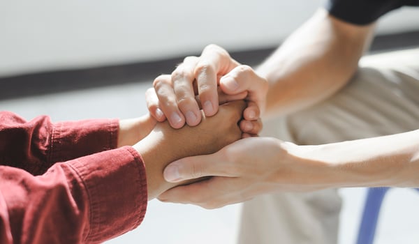 Close-up of two people holding hands in a gesture of comfort, one wearing a red shirt