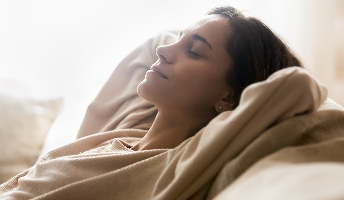 Person rests on a couch with eyes closed, head supported by pillows and covered with a blanket in soft natural light.