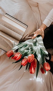 Overhead view of a hand holding red tulips on a bed beside an open book