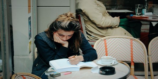 Young woman resting her chin on her hand while writing in a book at a sidewalk cafe table