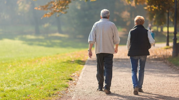 Two older adults walk side by side along a sunlit park path lined with autumn leaves, seen from behind