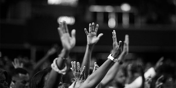 Crowd raising open hands at a dimly lit indoor gathering, in black and white
