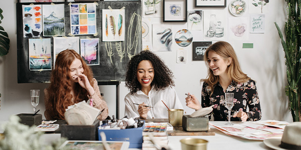 Three young women laughing together while painting with watercolors at a studio table