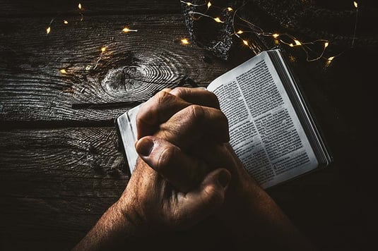 Praying hands over Bible surrounded by string lights