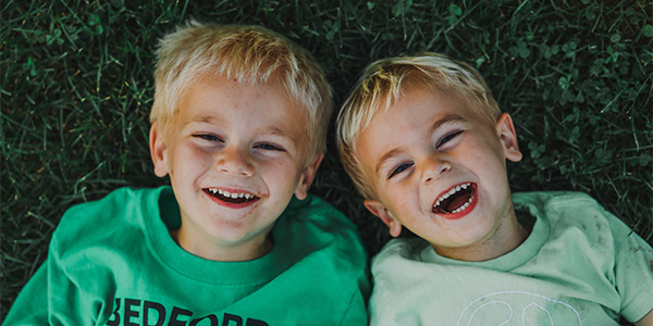 Two young blond boys in green T-shirts lie side by side in the grass, laughing