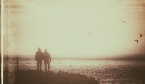 Two people stand side by side on seaside rocks as gulls fly overhead, in faded vintage sepia tones