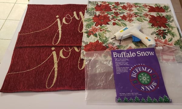 Holiday craft supplies: red fabric with gold joy lettering, poinsettia fabric, a hot glue gun and a bag of artificial snow