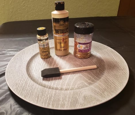 Supplies for upgrading a charger plate: gold metallic paint, gold glitter paint, a jar of glitter and a foam brush