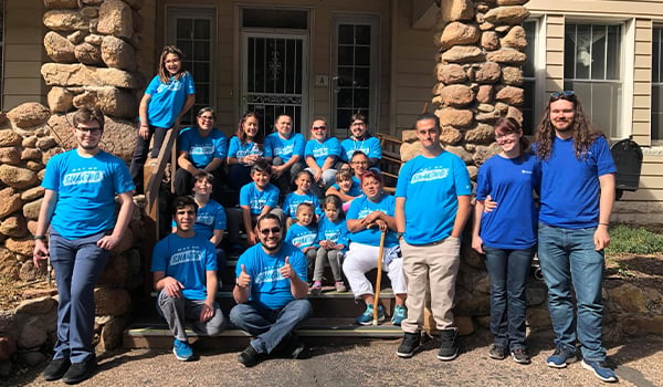 Adults and children in matching blue Day of Sharing T-shirts gather on the front steps of a stone-pillared house