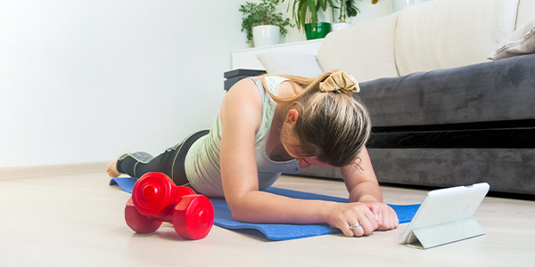 Woman holding a forearm plank on a yoga mat in a living room, with a tablet and red dumbbells nearby