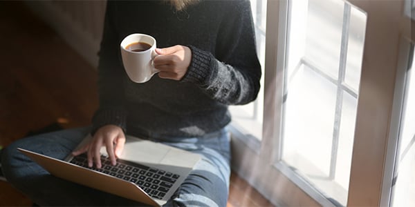 Person sitting on the floor by a window with a laptop on their lap and a cup of coffee in hand