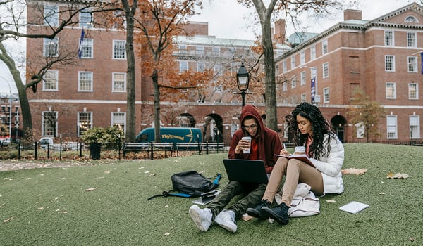 Two young adults study on a campus lawn with a laptop and notebook, brick buildings and autumn trees behind them