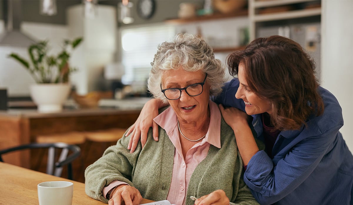 An older mom being hugged from behind by adult daughter.
