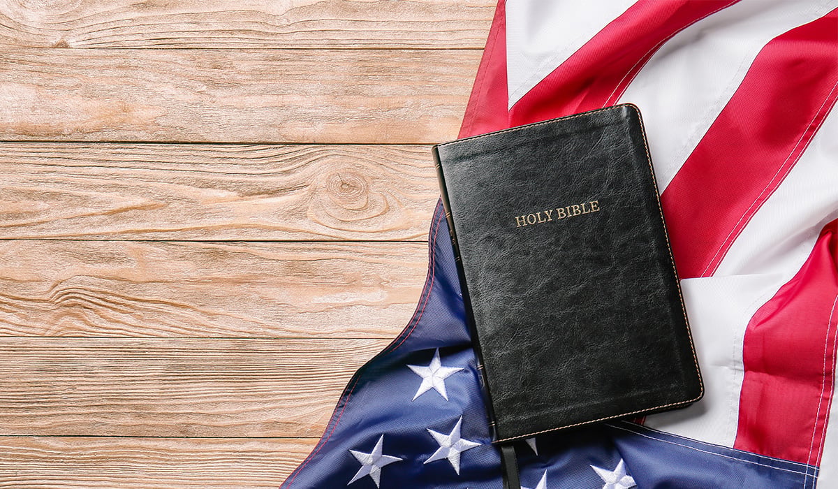 A bible and an American flag laying flat on a wood table.